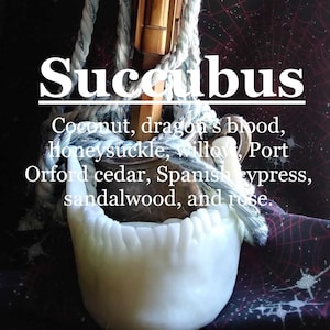May include: A white ceramic sculpture of a set of teeth, with the text "Succubus" above it. The text below the sculpture reads "Coconut, dragon's blood, honeysuckle, willow, Port Orford cedar, Spanish cypress, sandalwood, and rose."