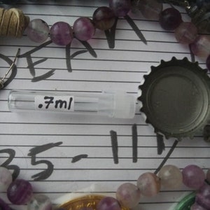 May include: A clear glass bottle with a black label that says ".7ml" is surrounded by purple and green beads. The bottle is lying on a white surface with black lines.