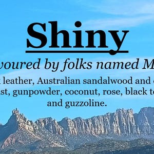 May include: A graphic design with the word "Shiny" in bold black text with a line underneath. The text "Favoured by folks named Max" is below the word "Shiny". Below that is a list of words: "Black leather, Australian sandalwood and desert dust, gunpowder, coconut, rose, black tea, and guzzoline."