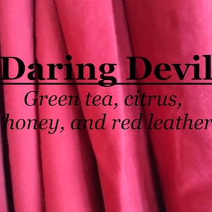 May include: A close-up of a pink fabric with the text "Daring Devil" and the words "Green tea, citrus, honey, and red leather."