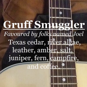 May include: A close-up of an acoustic guitar with a brown leather guitar strap. The guitar has a natural wood finish and a white text overlay that reads "Gruff Smuggler Favoured by folks named Joel Texas cedar, river algae, leather, amber, salt, juniper, fern, campfire, and coffee."
