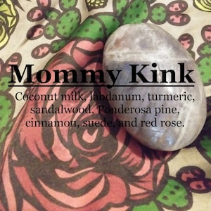 May include: A close-up of a smooth, gray and white stone with a floral pattern background. The text "Mommy Kink" is written in black letters above the stone. The text below the stone reads "Coconut milk, labdanum, turmeric, sandalwood, Ponderosa pine, cinnamon, suede, and red rose."