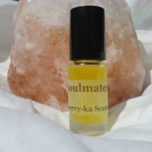 May include: A small glass bottle with a black cap and a yellow liquid inside. The bottle has a white label with the text "Soulmates" and "Berry-ka Scent" printed on it.
