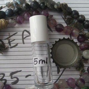 May include: A clear glass bottle with a white cap and a label that says "5 ml". The bottle is sitting on a surface with a variety of colorful beads.