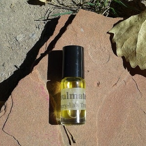 May include: A small, clear glass bottle with a black cap and a yellow label that reads "Soulmate" and "Berry-la's Tru". The bottle is sitting on a brown, textured surface.