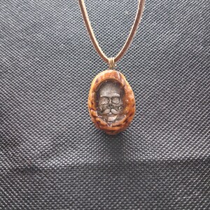 Puede incluir: Un collar con colgante marrón con un diseño de calavera tallada. La calavera tiene cuencas de los ojos, una nariz y un bigote. El colgante cuelga de un collar de cordón marrón. El colgante está enmarcado en un marco ovalado marrón.
