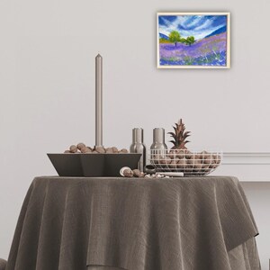 May include: An oil painting of a lavender field with green trees under a blue sky with white clouds. The painting is in a light wood frame. The painting is displayed on a table with a gray tablecloth, a black bowl, and a wire basket.