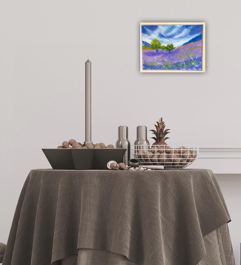 May include: An oil painting of a lavender field with green trees under a blue sky with white clouds. The painting is in a light wood frame. The painting is displayed on a table with a gray tablecloth, a black bowl, and a wire basket.