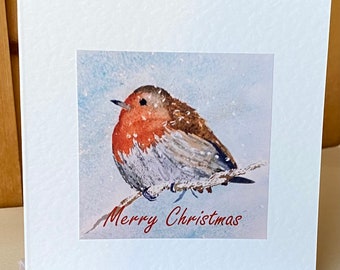 May include: A white greeting card featuring a watercolor painting of a robin perched on a branch in a snowy scene. The words "Merry Christmas" are written in red script below the image. The card is blank inside.