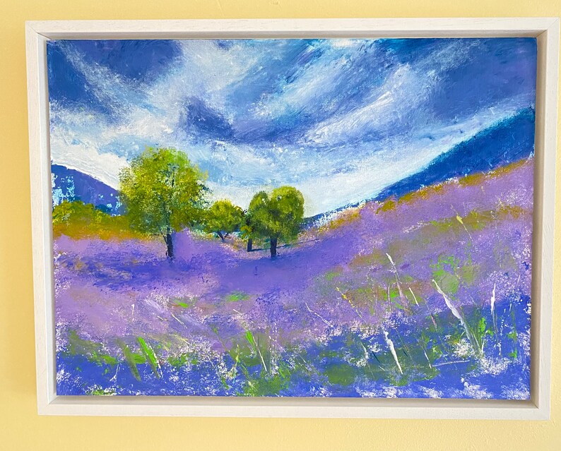 May include: An original landscape painting in a white frame. The artwork depicts a field of lavender flowers under a cloudy blue sky. Green trees dot the landscape, adding contrast to the purple and blue hues.
