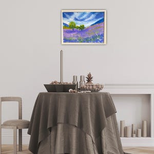 May include: An oil painting in a gold frame depicts a lavender field under a blue sky with white clouds. Two green trees stand on the horizon. The painting hangs above a table with a gray tablecloth, a candle, and decorative items.
