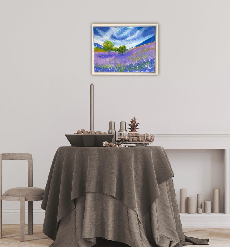 May include: An oil painting in a gold frame depicts a lavender field under a blue sky with white clouds. Two green trees stand on the horizon. The painting hangs above a table with a gray tablecloth, a candle, and decorative items.