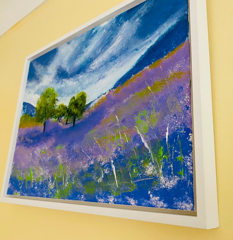 May include: An abstract landscape painting in a white frame. The artwork depicts a hillside covered in purple flowers, with green trees and a blue sky with white clouds. The painting uses thick brushstrokes and a vibrant color palette.