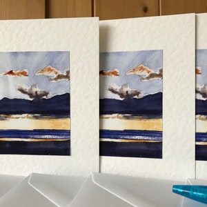 K&ouml;nnte beinhalten: Ein Set aus drei Aquarell-Landschaftsbildern. Jedes Gem&auml;lde zeigt eine dunkelblaue Bergkette, einen goldenen Horizont und ein blaues Gew&auml;sser unter einem bew&ouml;lkten Himmel. Die Gem&auml;lde sind in Wei&szlig; mattiert.