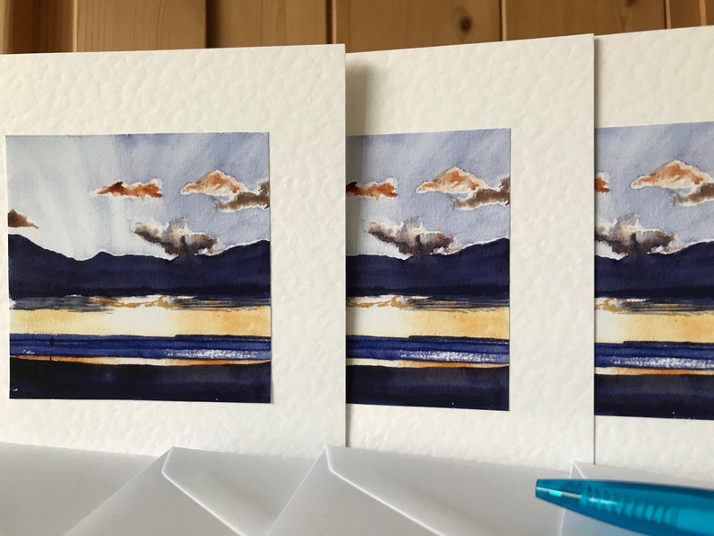 K&ouml;nnte beinhalten: Ein Set aus drei Aquarell-Landschaftsbildern. Jedes Gem&auml;lde zeigt eine dunkelblaue Bergkette, einen goldenen Horizont und ein blaues Gew&auml;sser unter einem bew&ouml;lkten Himmel. Die Gem&auml;lde sind in Wei&szlig; mattiert.