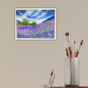 May include: Framed landscape painting depicting a field of purple flowers under a blue sky with white clouds. Green trees are in the middle ground. The painting is in a white frame.