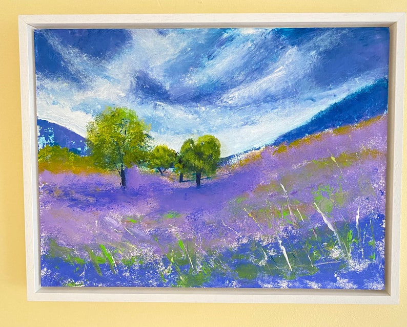 May include: An original landscape painting in a white frame. The artwork depicts a field of lavender flowers under a cloudy blue sky. Green trees and grass add contrast to the purple and blue hues. The painting evokes a sense of tranquility.
