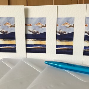 K&ouml;nnte beinhalten: Vier Aquarell-Landschaftsbilder mit einem Sonnenuntergang &uuml;ber dunklen Bergen und einem Gew&auml;sser. Die Gem&auml;lde sind in wei&szlig;en Passepartouts gerahmt und werden mit wei&szlig;en Umschl&auml;gen geliefert. Ein blauer Stift liegt auf den Umschl&auml;gen.