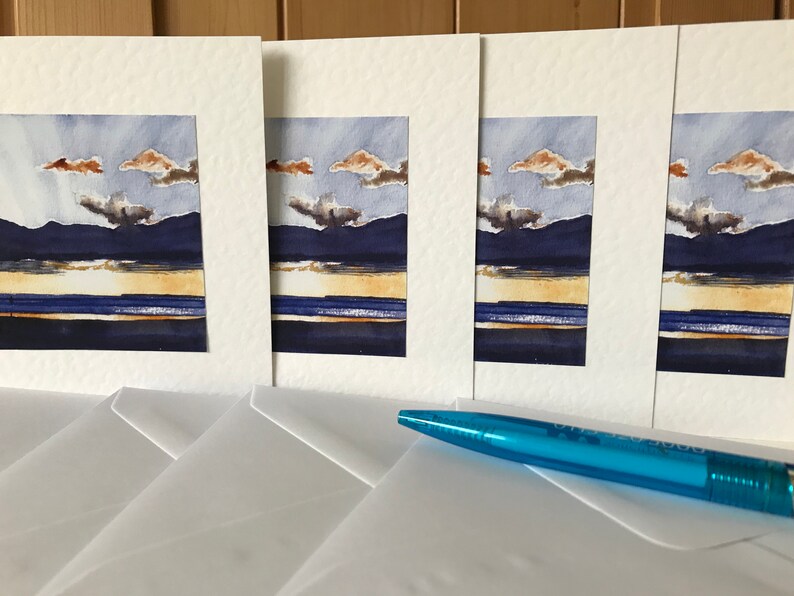 K&ouml;nnte beinhalten: Vier Aquarell-Landschaftsbilder mit einem Sonnenuntergang &uuml;ber dunklen Bergen und einem Gew&auml;sser. Die Gem&auml;lde sind in wei&szlig;en Passepartouts gerahmt und werden mit wei&szlig;en Umschl&auml;gen geliefert. Ein blauer Stift liegt auf den Umschl&auml;gen.