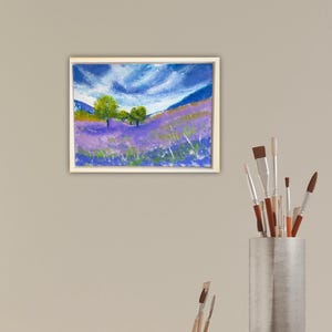 May include: A framed pastel painting depicts a lavender field under a cloudy blue sky. Green trees dot the landscape, with hints of yellow and green grass. The artwork is in a white frame.
