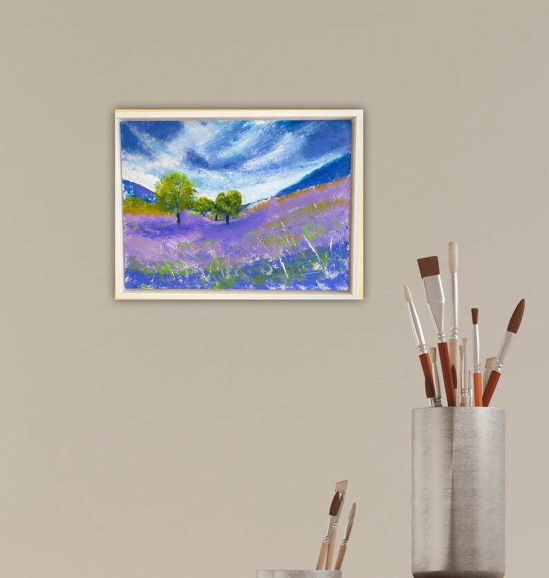 May include: A framed pastel painting depicts a lavender field under a cloudy blue sky. Green trees dot the landscape, with hints of yellow and green grass. The artwork is in a white frame.