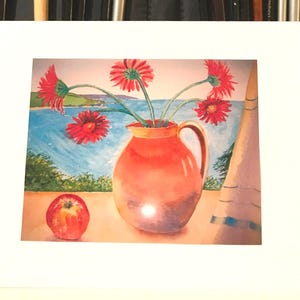 May include: Watercolor painting of a still life featuring a red pitcher with red flowers, a red apple, and a coastal scene in the background. The painting is framed in a white mat.