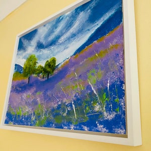 May include: An abstract landscape painting in a white frame. The artwork depicts a hillside covered in purple flowers, green trees, and a blue sky with white clouds. The painting has a textured, impressionistic style.
