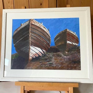 May include: A framed painting depicting two weathered wooden boats resting on a brown, textured shore against a vibrant blue sky. The boats feature brown planks with white painted sections at the bow, captured in a realistic style within a white frame.