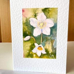 May include: A white greeting card with a watercolor painting of white flowers with yellow centers against a green and yellow background. The card has a textured, white border.