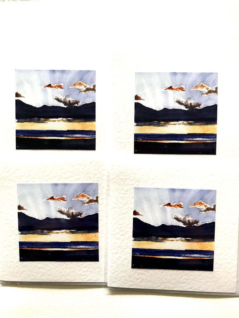 Four blank greeting cards with watercolor paintings of a sunset over a mountain range and a body of water. The cards are white with a textured surface. The paintings are in shades of blue, purple, brown, and yellow.