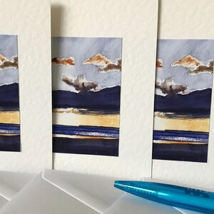 Four white cards with blue, yellow, and brown watercolor paintings of a landscape with mountains and clouds. The cards are stacked on top of each other and are surrounded by white envelopes.