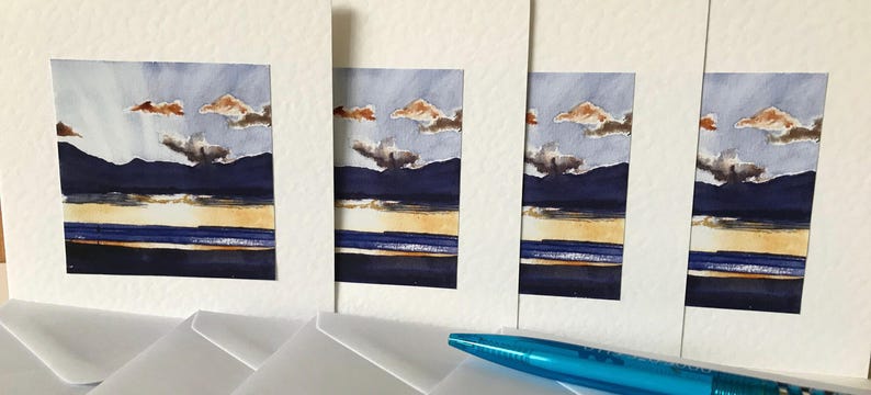 Four white cards with blue, yellow, and brown watercolor paintings of a landscape with mountains and clouds. The cards are stacked on top of each other and are surrounded by white envelopes.