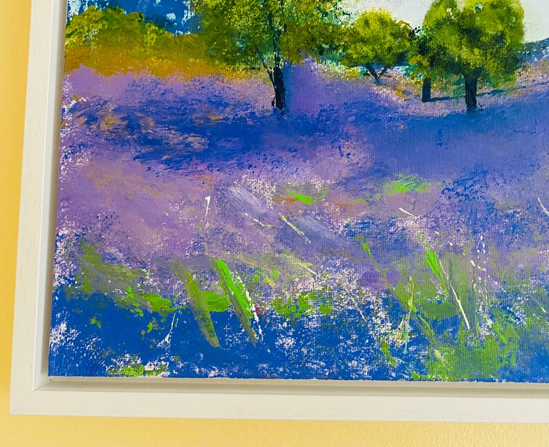 May include: An original acrylic painting of a lavender field with trees under a blue sky. The artwork is framed in a white wooden frame. The painting features shades of purple, blue, green, and white, creating a vibrant landscape scene.
