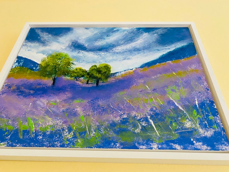 May include: An original landscape painting in a white frame. The artwork depicts a field of purple flowers under a blue sky with white clouds. Green trees and grass are also visible, creating a serene and colorful scene.