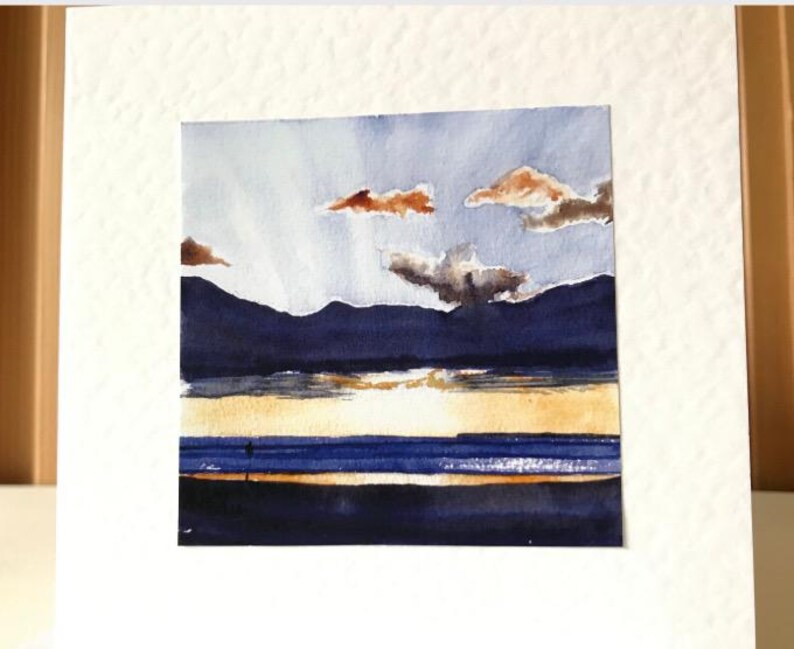K&ouml;nnte beinhalten: Aquarellmalerei einer Landschaft mit Sonnenuntergang &uuml;ber dem Wasser. Der Himmel ist hellblau mit orangefarbenen und grauen Wolken. Dunkelblaue Berge befinden sich im Hintergrund, und das Wasser reflektiert die Farben des Sonnenuntergangs.