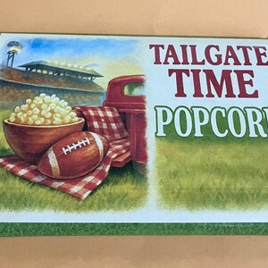 Może przedstawiać: Kolorowa ilustracja sceny z tailgate'u z piłką nożną, popcornem i czerwoną ciężarówką. Słowa "TAILGATE TIME POPCORN" są wyświetlane pogrubioną czcionką. Tło przedstawia stadion i zieloną trawę.