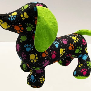 May include: A black plush dachshund toy with a green ear and tail. The toy is covered in a colorful paw print pattern.