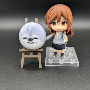 Pode incluir: Uma pequena figura em estilo anime com cabelo castanho, um suéter cinza e uma saia preta está ao lado de um bicho de pelúcia azul claro em um cavalete em miniatura. O bicho de pelúcia tem um rosto sorridente e detalhes azuis.