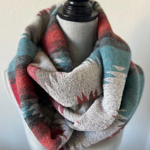 May include: A gray, red, and teal infinity scarf with a geometric pattern. The scarf is made of a woven fabric and is draped over a mannequin head. The design includes triangles and stripes.