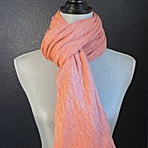 May include: A pink cable knit scarf draped over a mannequin. The scarf is a soft, light pink color and has a classic cable knit pattern.