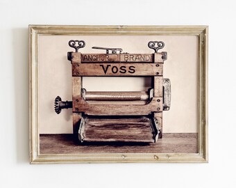 May include: An antique wooden clothes wringer with the text "Anchor Brand Voss" in a framed print. The wringer has a crank, rollers, and a tray. The print is in a gold-colored frame.