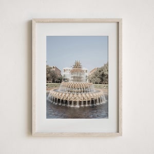 May include: Framed print featuring a tiered fountain with water cascading into a pool. The fountain is centered in the image, with buildings and trees in the background under a clear blue sky. The frame is light-colored.