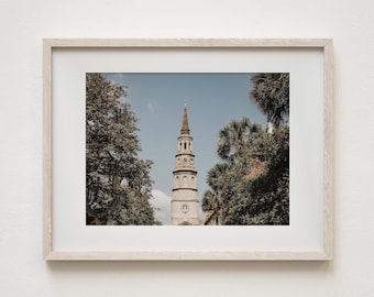 May include: Framed print of a tall, white church steeple against a blue sky, framed by lush green trees. The steeple has multiple tiers and a pointed top. The print is in a light-colored wooden frame.