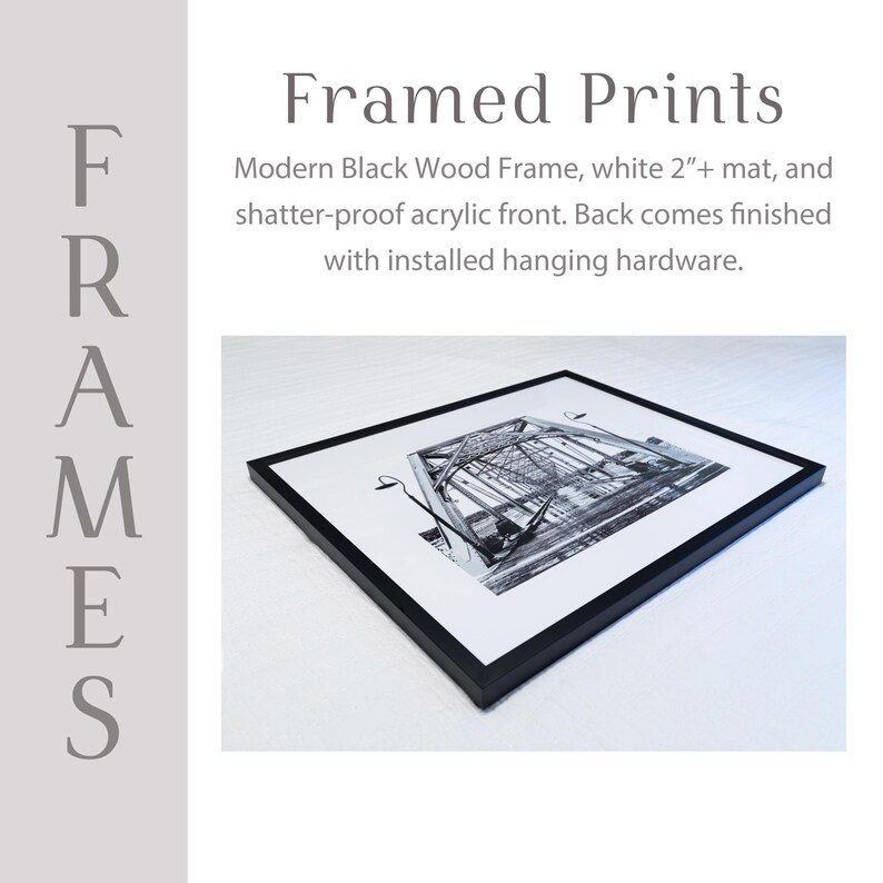 The image is a framed print featuring a black and white photograph of a bridge. The print is placed on a white surface and has a modern black wood frame. The back of the print includes a white mat and a shatter-proof acrylic front. The print is finished with installed hanging hardware.