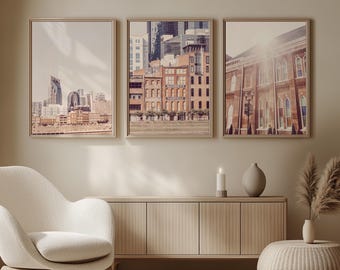 May include: Three framed art prints featuring cityscapes in warm tones. The prints depict buildings and skylines, with a neutral color palette. The frames are gold-toned, and the artwork is displayed above a wooden cabinet and a white chair.
