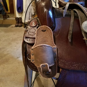 Può includere: Una sella in pelle marrone con un design lavorato e un portautensili in pelle marrone attaccato al lato. Il portautensili ha una patta con chiusura a scatto e una fibbia in metallo.