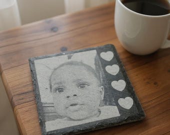 May include: A square slate coaster featuring a black and white image of a baby's face. White heart shapes are on the right side of the coaster. A white mug filled with a dark liquid sits on a wooden table.