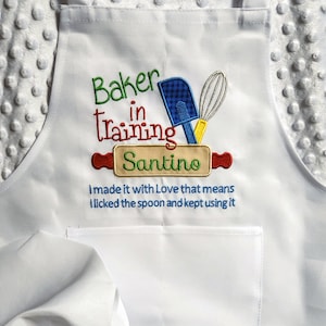 May include: White apron with embroidered text "Baker in Training Santino" and a blue spatula, yellow whisk, and red rolling pin. The text below reads "I made it with Love that means I licked the spoon and kept using it."
