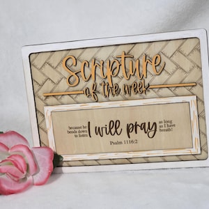 May include: Wooden sign with the text "Scripture of the week" in a decorative font. The sign also includes the phrase "I will pray" and a Bible verse. A pink and white rose is in the foreground.