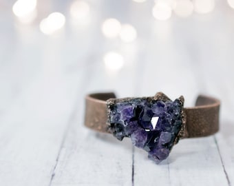 May include: A copper-colored cuff bracelet featuring a large, raw amethyst crystal. The deep purple crystal is the focal point, set against the textured metal band. The bracelet is displayed on a light-colored wooden surface.